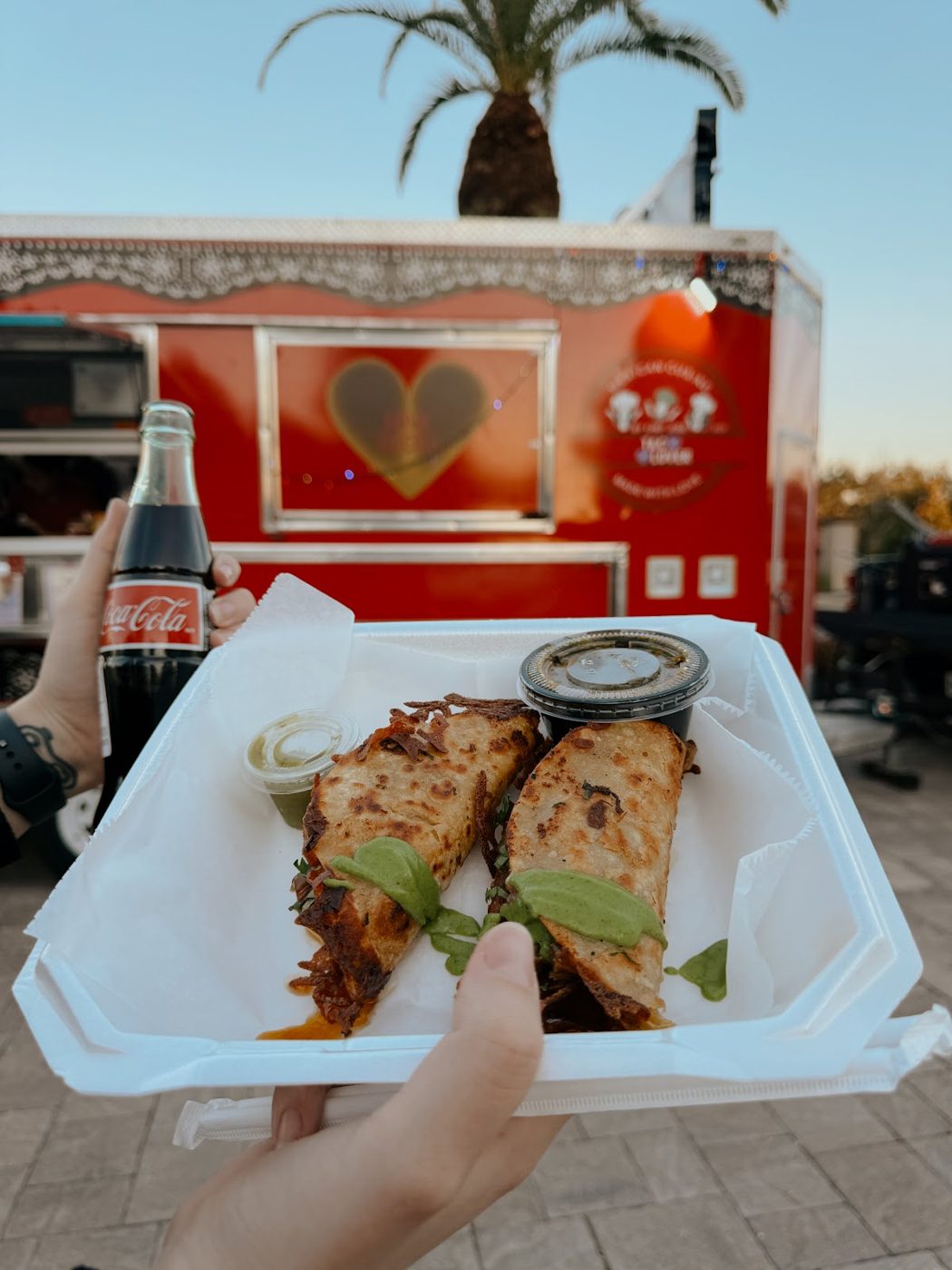 Crispy birria quesadilla tacos with avocado salsa held in front of the red Tacolover truck at golden hour